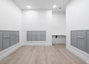 Thumbnail 19 of 29 - A room with white walls and wooden flooring, featuring a row of grey lockers on the left and a bench on the right.