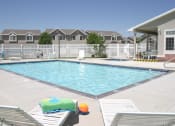 Thumbnail 3 of 3 - View of the blue swimming pool with a white fence, white chaise lounge chairs with a colorful towel on one and a multicolored beach ball in between, and resident building to the right at Oakstone Apar