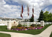 Thumbnail 2 of 3 - View of the front of Oakstone Apartments with property signage, three flags, and manicured brightly-colored flowers and walking pathways through the green grass at Oakstone Apartments in Utah 84015
