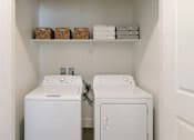 Thumbnail 28 of 28 - A white laundry room with two washing machines and a shelf with baskets and towels.