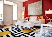 Thumbnail 1 of 17 - A white sofa with red and orange pillows sits in a room with a red wall and a white chair.