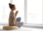 Thumbnail 6 of 30 - A woman in a beige sweater is sitting on a woven stool by a window.