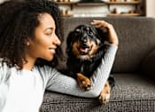 Thumbnail 16 of 20 - A woman with curly hair is sitting on a grey couch with a black and tan dog on her lap.