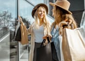 Thumbnail 8 of 30 - Two women wearing hats and carrying shopping bags are standing outside a store.