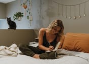 Thumbnail 3 of 26 - a woman sitting on a bed reading a book with a cat in the background