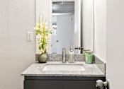 Thumbnail 9 of 29 - A bathroom sink with a green jar and a vase with white flowers.