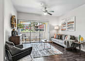 Thumbnail 4 of 64 - A living room with a grey couch, a black chair, a wooden side table, a floor lamp, a ceiling fan, and a large window with a view of the outside.