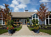 Thumbnail 1 of 34 - A house with a grey roof and a white door is surrounded by trees and flower pots.