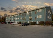 Thumbnail 1 of 13 - A parking lot in front of a grey apartment building with cars parked.