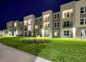 Thumbnail 41 of 62 - Apartment complex with green lawn and lights on.