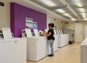 Thumbnail 8 of 18 - a woman standing at a line of washing machines in a laundromat at Hamilton Manor Apartments, Hyattsville, MD