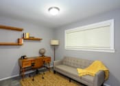 Thumbnail 22 of 81 - A room with a grey couch, a desk with a chair, a lamp, and a window with blinds.