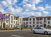 Thumbnail 1 of 7 - A row of apartment buildings with people walking in front.