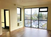 Thumbnail 20 of 26 - A room with wooden floors and a view of a parking lot through the windows.
