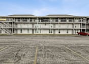 Thumbnail 9 of 25 - A parking lot in front of a white building with a red car parked in the lot.