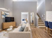 Thumbnail 6 of 18 - A living room with a beige couch, a coffee table, and a staircase.
