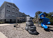Thumbnail 12 of 15 - A playground with a blue slide and grey sandbox in front of a grey building.