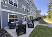 Thumbnail 9 of 15 - A building with a grey facade has a patio with black chairs and tables.