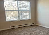 Thumbnail 6 of 21 - A room with a carpeted floor, a window with blinds, and a wall-mounted heater.