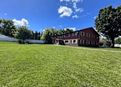 Thumbnail 3 of 4 - A large grassy field with a white building and a red building in the background.