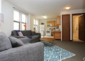 Thumbnail 3 of 25 - A grey couch with a blue throw pillow sits in a living room with a kitchen in the background.