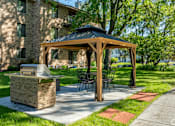 Thumbnail 20 of 25 - A wooden gazebo with a grill is situated in a grassy area in front of a building.
