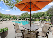 Thumbnail 22 of 38 - Poolside patio dining with umbrella and seating at Parks on the Green Apartments, Temple, TX 76504