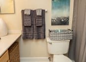 Thumbnail 10 of 27 - A bathroom with a toilet, a mirror, a towel rack with two towels, a basket, and a painting on the wall.at Deer Park Apartments, Randallstown, 21133