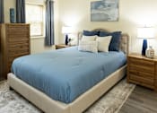 Thumbnail 13 of 22 - A bed with a blue cover sits in a room with a wooden dresser and a painting on the wall.