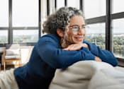 Thumbnail 10 of 33 - A person with curly hair wearing glasses and a blue top is sitting on a couch.