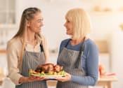 Thumbnail 6 of 8 - Two women in aprons holding a plate with a roasted turkey.