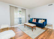 Thumbnail 3 of 41 - Chattanooga, TN, Apartments for Rent - Riverview North - Spacious Living Room with Wood-Style Flooring, Area Rug, Blue Sofa, Cushioned Bench/Coffee Table, White Walls, Fluffy Stools, Side Table, Floor Lamp, and Sliding Glass Door with Access to Patio.