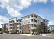 Thumbnail 45 of 46 - Pet-Friendly Apartments in Norcross - The Darnell - Building Exterior with Streetview, with Parking Spots, Brick Details, and White Balconies Visible
