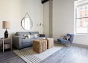 Thumbnail 8 of 25 - A living room with a grey couch, a round mirror, a lamp, a chair, and a wicker ottoman.