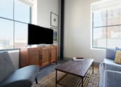 Thumbnail 9 of 25 - A living room with a grey couch, a wooden coffee table, and a black television.