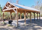 Thumbnail 27 of 27 - A wooden pavilion with a metal roof is surrounded by trees.
