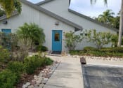 Thumbnail 3 of 34 - a white building with a blue door and a walkway with plants and trees at L'Estancia, Sarasota, 34231