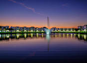 Thumbnail 55 of 78 - A fountain in the middle of a body of water with buildings in the background.
