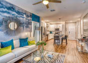 Thumbnail 3 of 34 - Apartments for Rent in Maitland, FL - The Parker at Maitland Station - Living Room with Wood Vinyl Plank Flooring, Area Rug, Couch, Standing Lamp, Glass-Top Coffee Table, Ceiling Fan, and View of Kitchen.