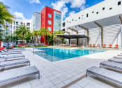 Thumbnail 18 of 34 - Maitland, FL Apartments - Parker at Maitland Station - Resort-Style Pool with Sundeck, Lounge Chairs, Covered Areas, Umbrellas, Tables and Chairs, Lawn Area with Red Seats, Surrounding Plants and Palm Trees, and View of Apartment Building