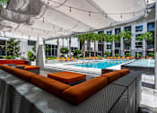 Thumbnail 14 of 39 - Pet-Friendly Luxury Apartments in Orlando, FL - NORA - An Outdoor Lounge Featuring Dark Wicker Couches, Ottomans, Orange Cushions, Canvas Canopy, String Lights, and View of the Pool Area.