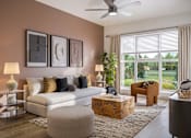 Thumbnail 12 of 16 - A living room with a white couch, a wooden coffee table, and a large window with blinds.