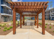 Thumbnail 22 of 28 - Dog-Friendly Apartments in Huntsville, AL - The Kelvin - Courtyard Grilling Station under Gazebo with Manicured Plants, Concrete Walkways, and View of Apartment Buildings.