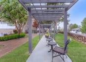Thumbnail 29 of 41 - Apartments in Chattanooga, TN - Riverview North - Outdoor Seating Area Under Pergola with Black Chairs, Tables, Manicured Grass Lawn, Trees, and Stone Wall.
