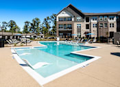 Thumbnail 5 of 45 - Cumming, GA, Luxury Apartments Near Alpharetta - Aura Creekside - Swimming Pool With In-Water Loungers, Pool Deck Seating, And Modern Apartment Buildings.
