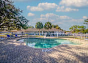 Thumbnail 1 of 15 - A pool surrounded by a brick patio with a white fence and trees in the background.