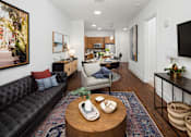 Thumbnail 7 of 57 - Decatur, GA, Apartments Near Marta Station - Arlo - Living Room With Black Sofa, Round Wood Coffee Table, Wall-Mounted Television, Dining Area, And Modern Kitchen In The Background.