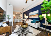 Thumbnail 7 of 58 - Buckhead, GA, Luxury Apartments Near Phipps Plaza- 92 West Paces - Furnished Living Room with Hardwood Flooring, Area Rug, Blue Accent Wall, White Sofa, Coffee Table, Side Tables with Lamps, Wooden Cabinet, Wall-Mounted TV, Houseplants, and View of Kitchen.