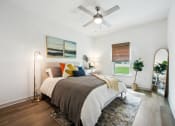Thumbnail 6 of 24 - Flagler Beach, FL, Luxury Apartments with In-Unit Washer and Dryer - Preserve at Flagler Beach - Bedroom with Wood-Inspired Flooring, Area Rug, Bed, Floor Lamp, Nightstand with Lamp, Standing Mirror, Ceiling Fan, and Window.