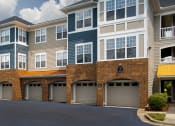Thumbnail 43 of 59 - Luxury Apartments in Chapel Hill, NC, with In-Unit Washers and Dryers - The Reserve at Meadowmont - Property Exterior with Blue and Tan Walls, Stone Details, White Trim, Beige Attached Garages, Large Windows, and Manicured Hedges.